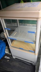 広島市内の店舗にて、小型の冷蔵ショーケースを運搬する便利屋万。ガラス面を毛布とパッドで厳重に保護し、台車で慎重に移動させている様子。