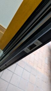 広島市内の住宅にて、閉まりにくくなった玄関ドアの建て付け調整を行う便利屋万の店主。ドアノブ（ラッチ）と鍵の噛み合わせをドライバーで精密に調整中。